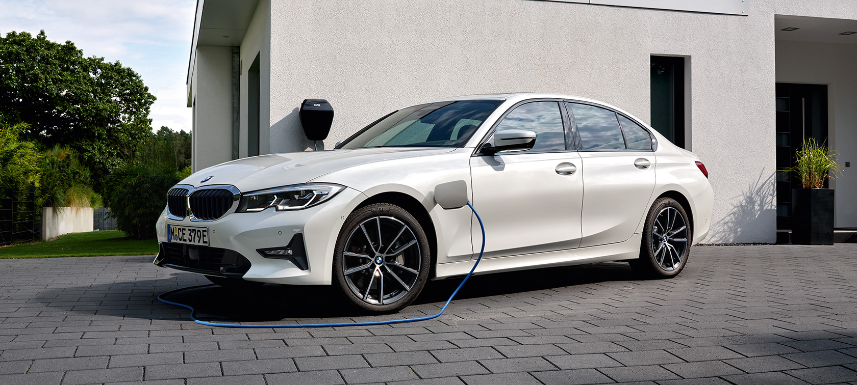 BMW Série 3 Berline hybride rechargeable 2021 G20, branchée à une borne de recharge, vue de trois quarts de profil BMW Série 3 Berline hybride rechargeable 2021 G20 devant un garage, vue de trois quarts de profil