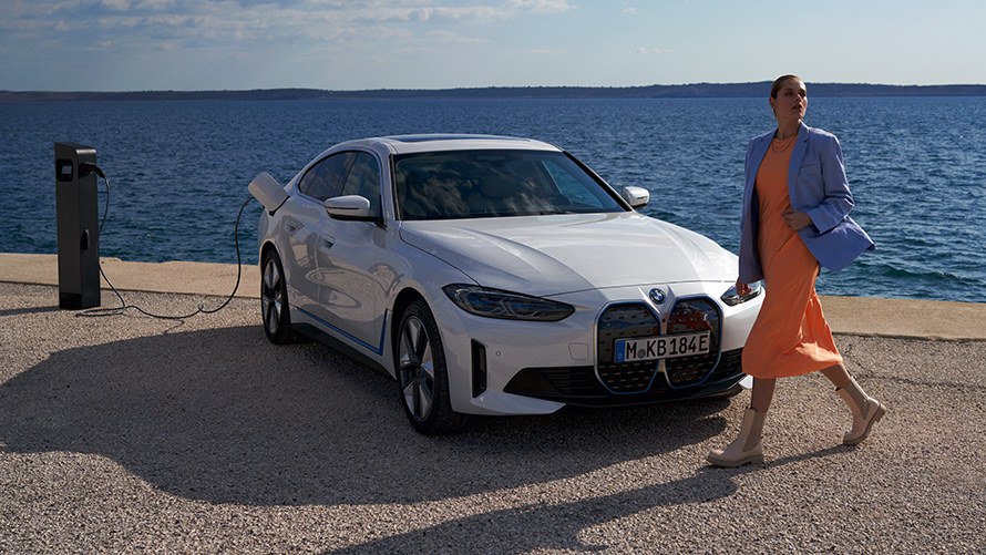 Vue de trois quarts avant de la BMW i4 eDrive40 G26 2021 à une borne de recharge Femme devant la BMW i4 eDrive40 G26 2021 Mineralweiß vue de trois quarts avant d'une borne de recharge devant un lac
