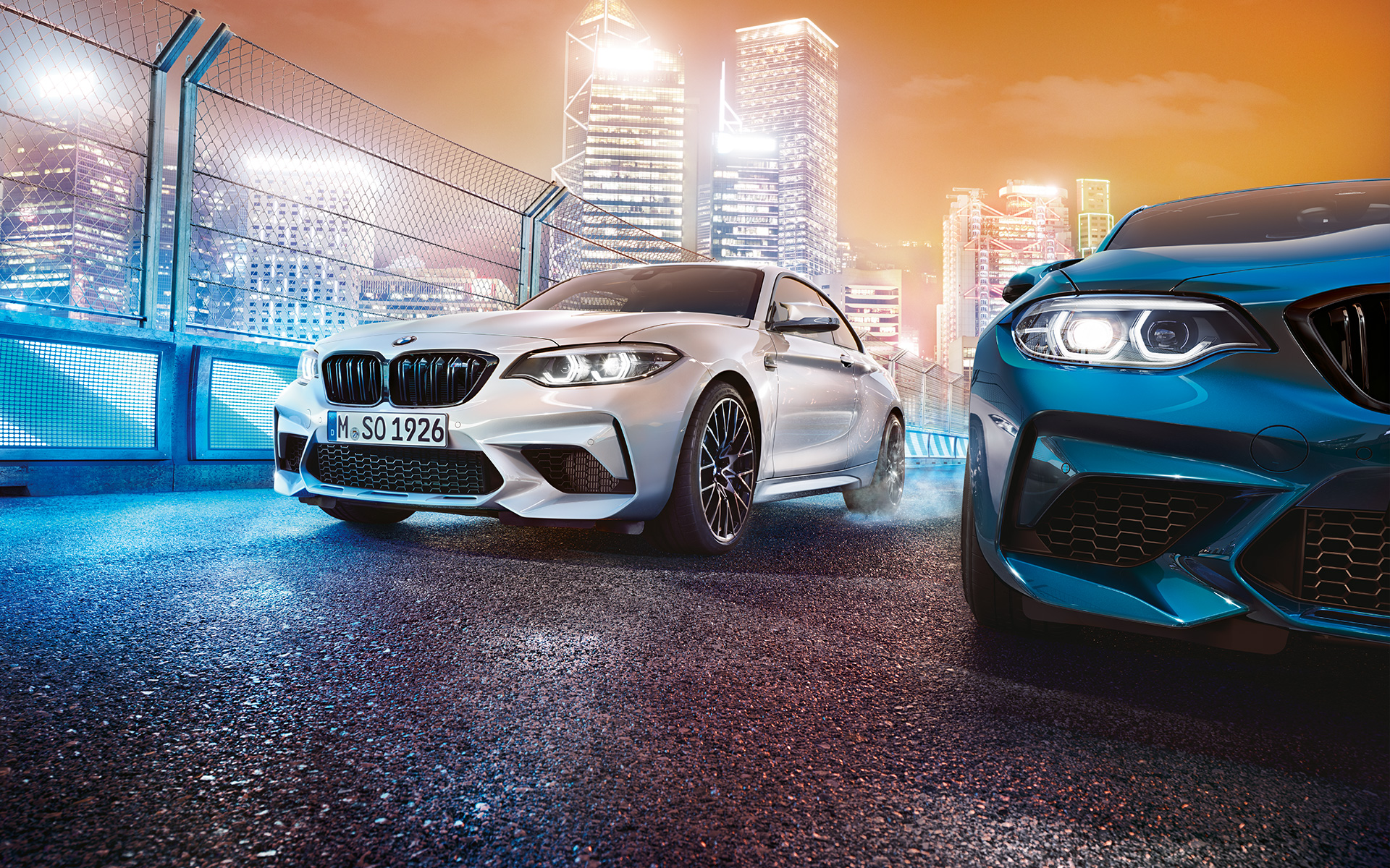 Vue avant BMW M2 Competition Une BMW M2 Competition blanche et une autre bleue vues de l'avant, dans le décor nocturne d'une grande ville.