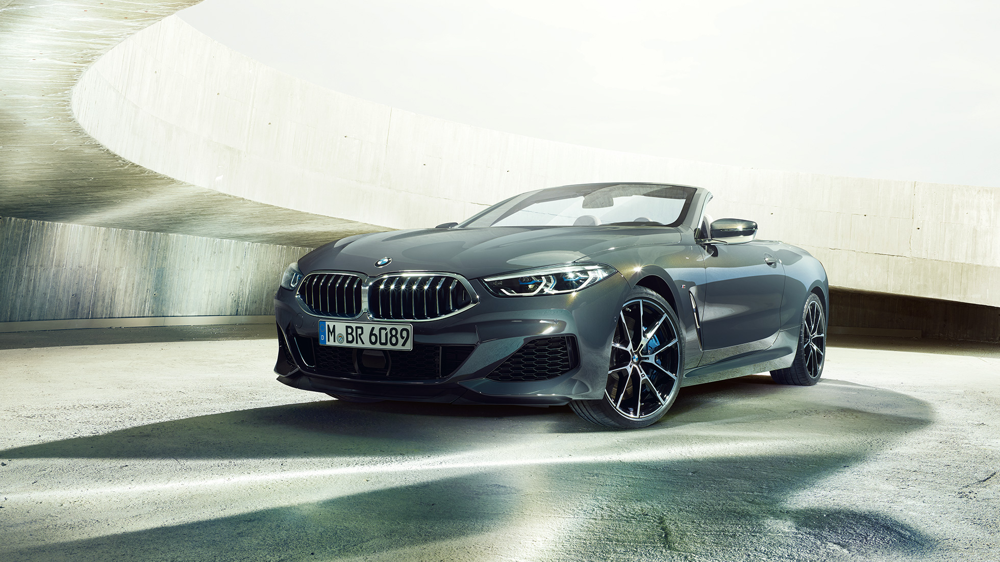 BMW M850i xDrive Cabriolet, vue de trois quarts avant G14. BMW M850i xDrive Cabriolet en Dravitgrau metallic avec capote ouverte, vue de trois quarts avant, à l’arrêt dans un bâtiment en béton G14.