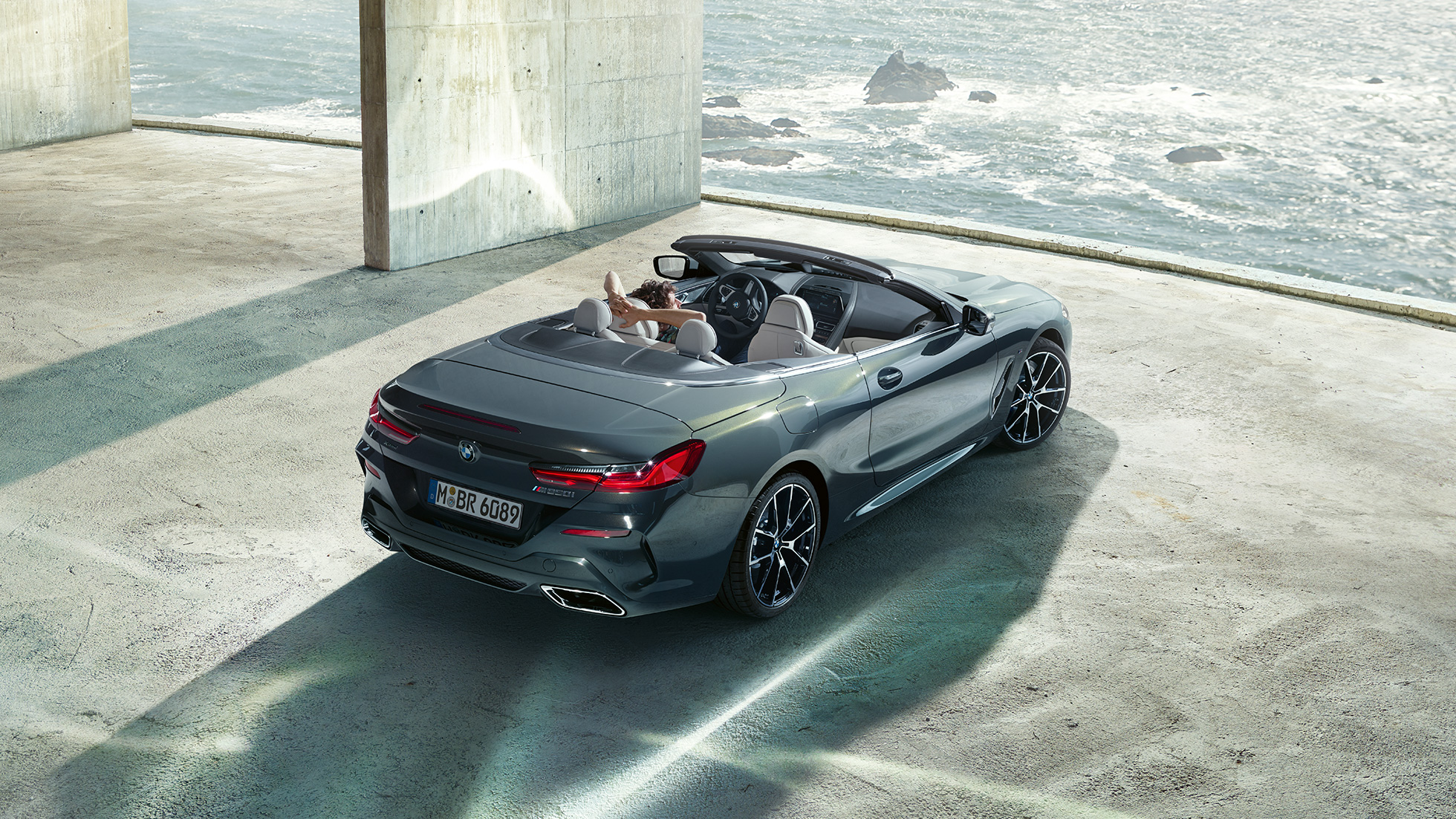 BMW M850i xDrive Cabriolet, vue en plongée G14. BMW M850i xDrive Cabriolet en Dravitgrau metallic avec capote ouverte, vue en plongée, en stationnement dans un bâtiment en béton devant l’océan G14.