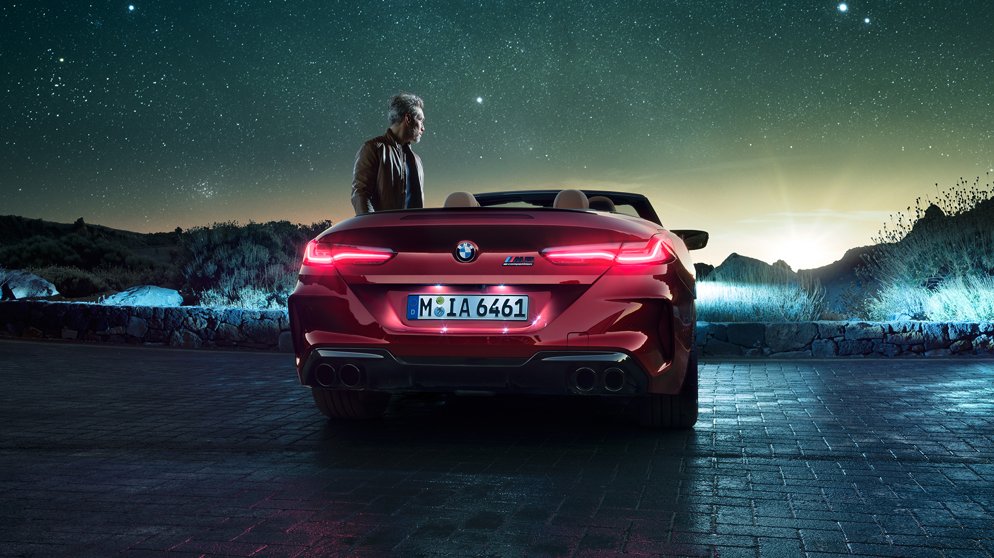 BMW M8 Competition Cabriolet, vue arrière F91. BMW M8 Competition Cabriolet en Motegi Rot metallic avec capote ouverte, vue arrière, à l’arrêt avec homme sous un ciel étoilé F91.