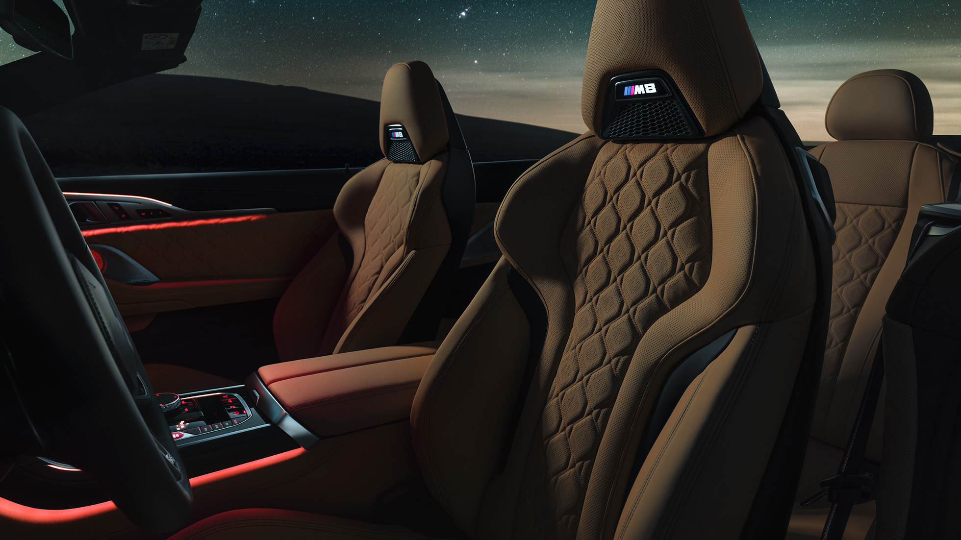 Siège M Sport en cuir Merino F91. BMW M8 Competition Cabriolet avec capote ouverte sous un ciel étoilé, habitacle avec sièges M sport en cuir Merino et éclairage d’ambiance rouge F91.