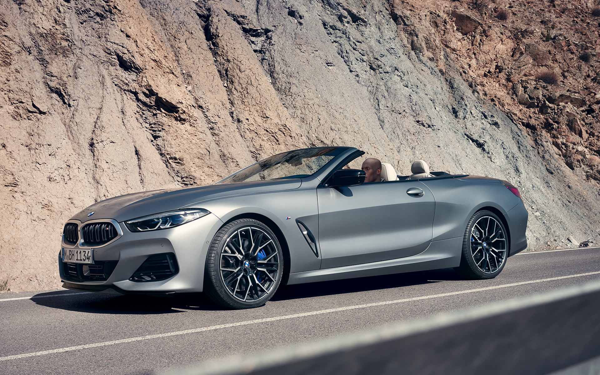 La nouvelle BMW M850i xDrive Cabriolet G14 LCI restylage 2022 BMW Individual Frozen Pure Grey metallic, vue de trois quarts avant en mouvement devant des roches