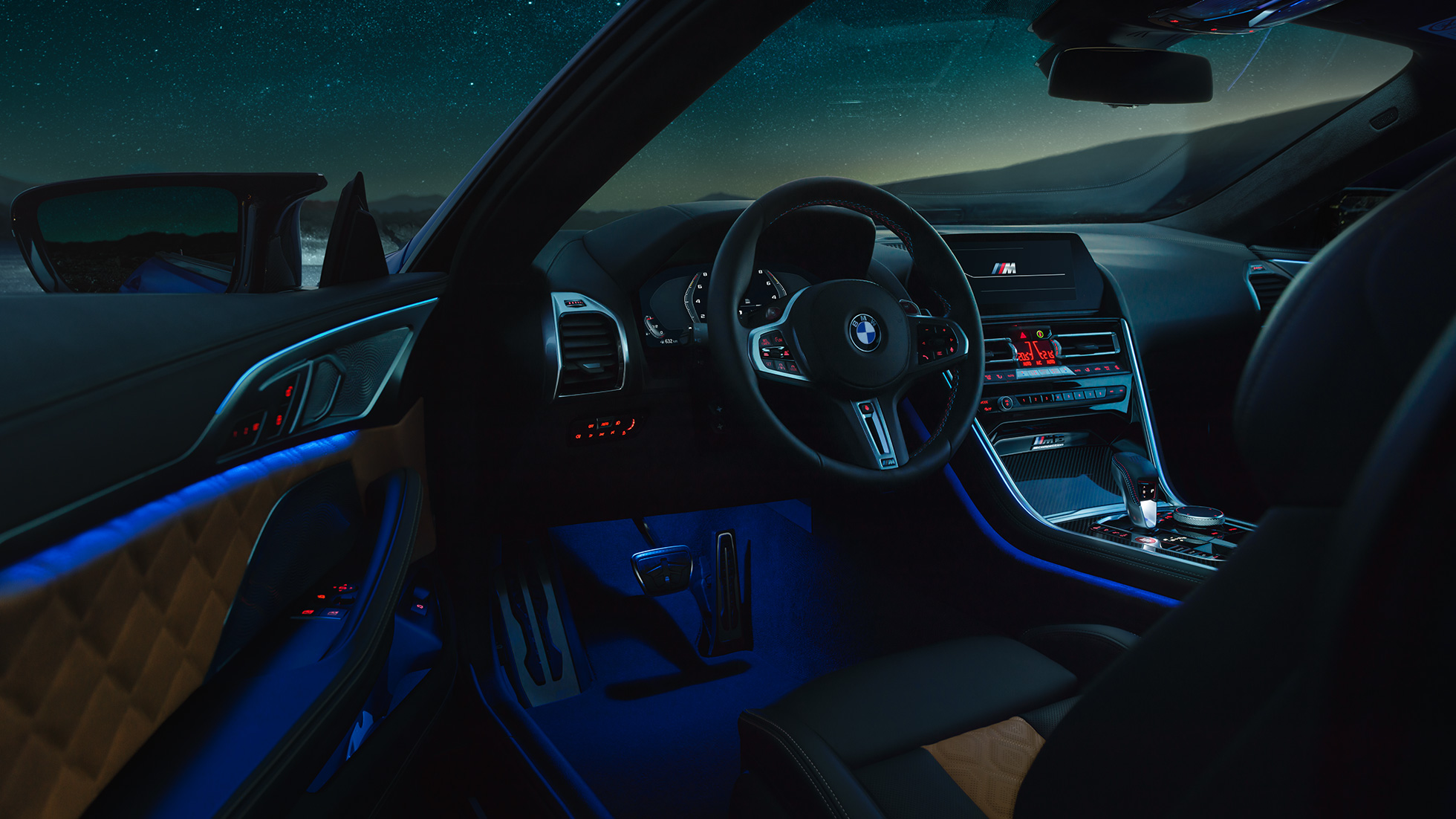 Intérieur de la BMW M8 Competition Coupé F92. BMW M8 Competition Coupé, habitacle vu de nuit, avec volant M en cuir et éclairage d’ambiance bleu, porte du conducteur ouverte F92.