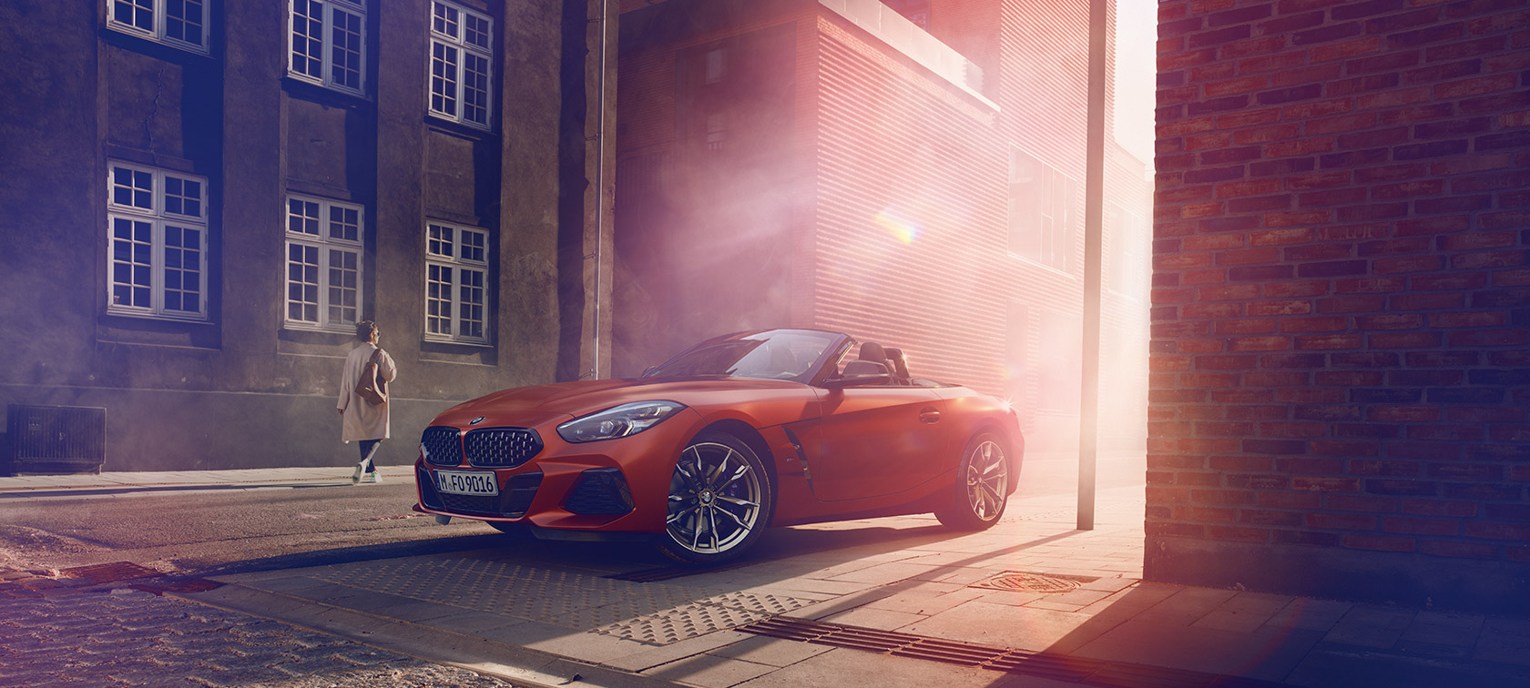 BMW Z4 First Edition orange dans la route BMW Z4 First Edition de couleur orange dans une rue plongée dans la pénombre devant un groupe de maisons. Une femme est présente.