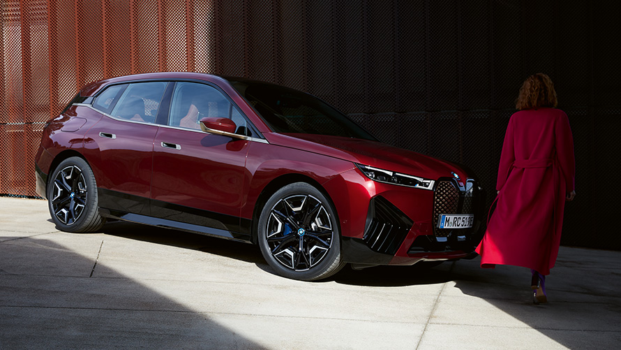 Femme devant la BMW iX i20 2021 vue de trois quarts face Femme avec manteau rouge devant la BMW iX i20 2021 vue de trois quarts face