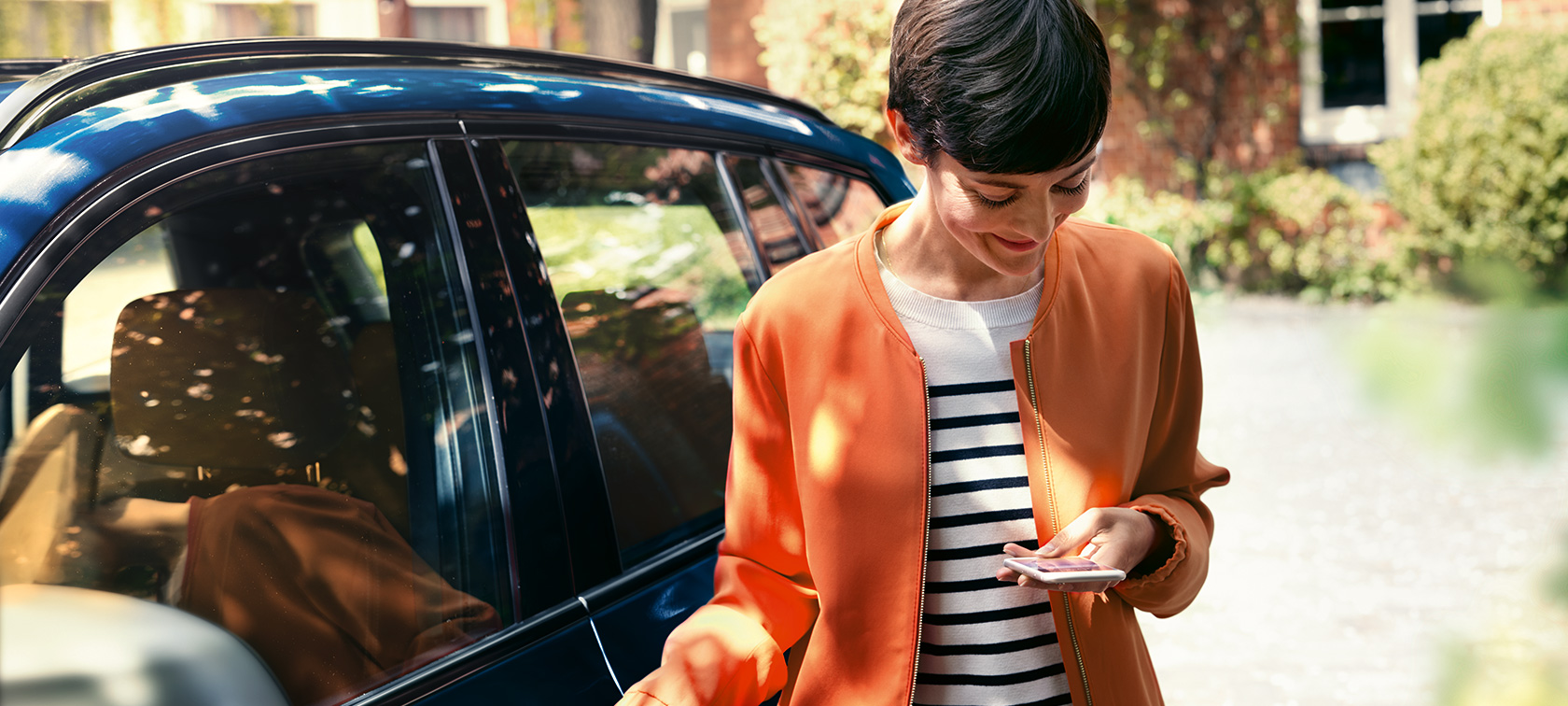 Femme aux cheveux sombres devant une BMW Femme aux cheveux sombres en blouson orange, devant une BMW, regardant son smartphone.