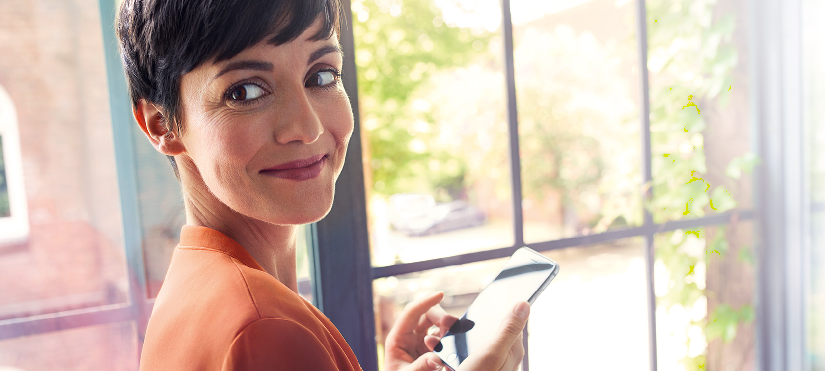 Femme aux cheveux sombres avec smartphone Femme aux cheveux sombres en blouson orange se tenant à la fenêtre avec son smartphone, elle sourit.