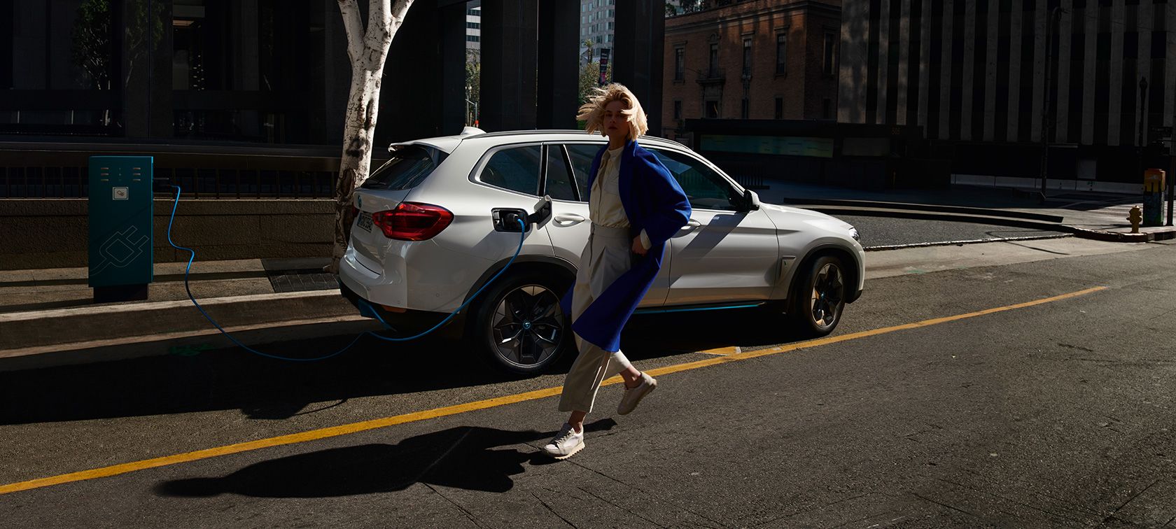 BMW iX3 en train de recharger à une borne dans la rue avec une passante BMW iX3 en train de recharger à une borne dans la rue avec une passante